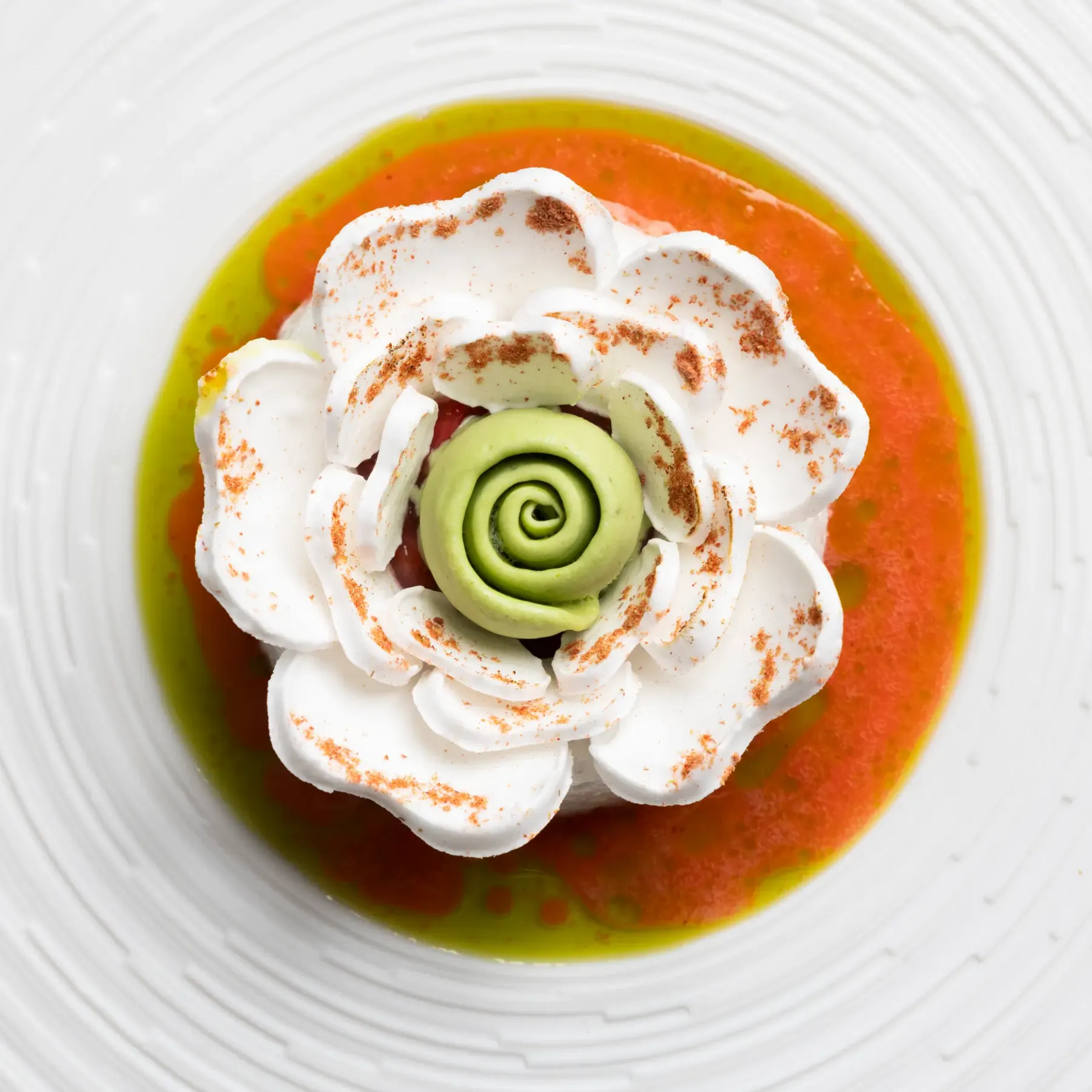 A dessert consisting of a white flower-shaped mousse, a green swirl, and an orange and green sauce on a white plate.