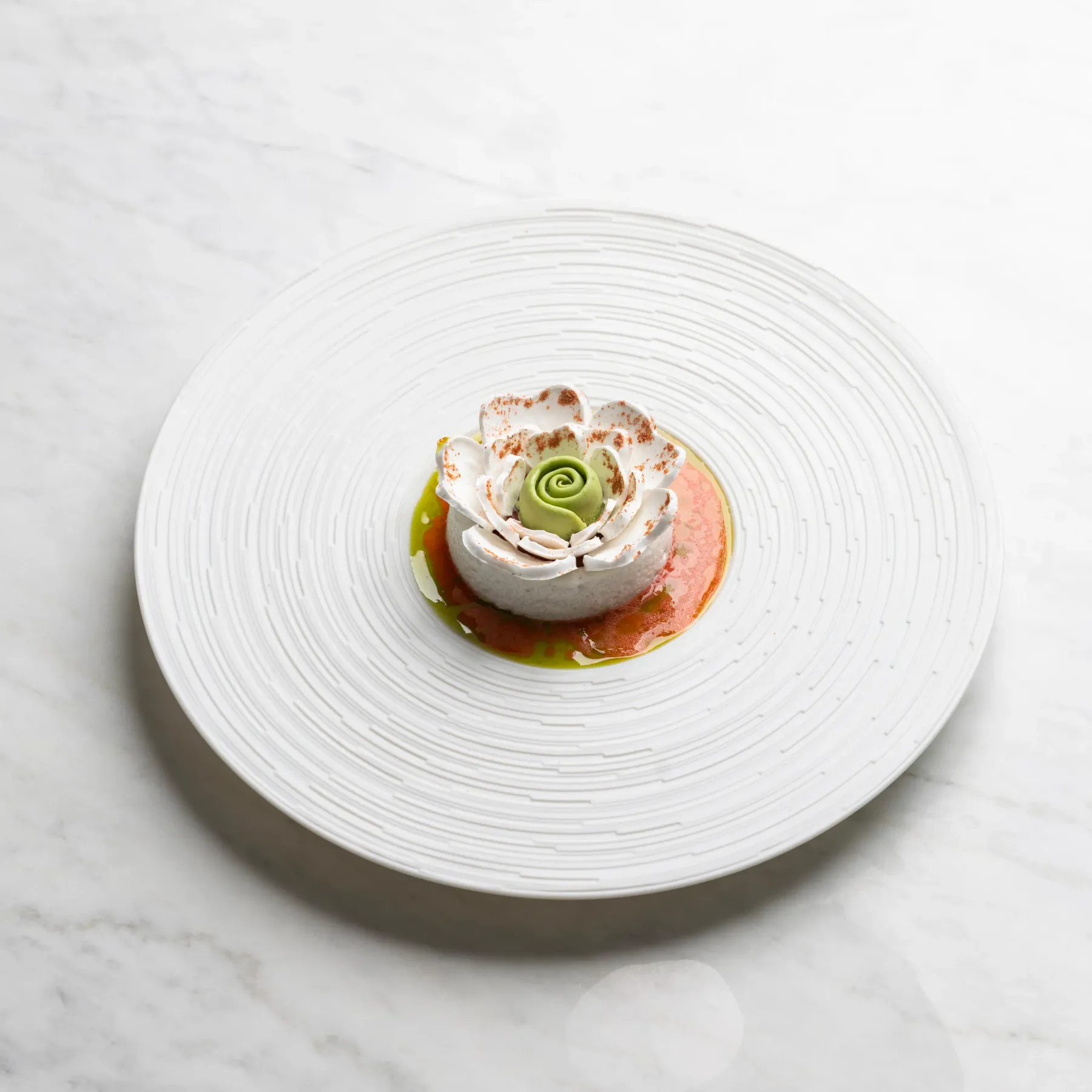 A small white rose-shaped dessert with a green touch at the center, placed on a circular white plate on a white marble surface.