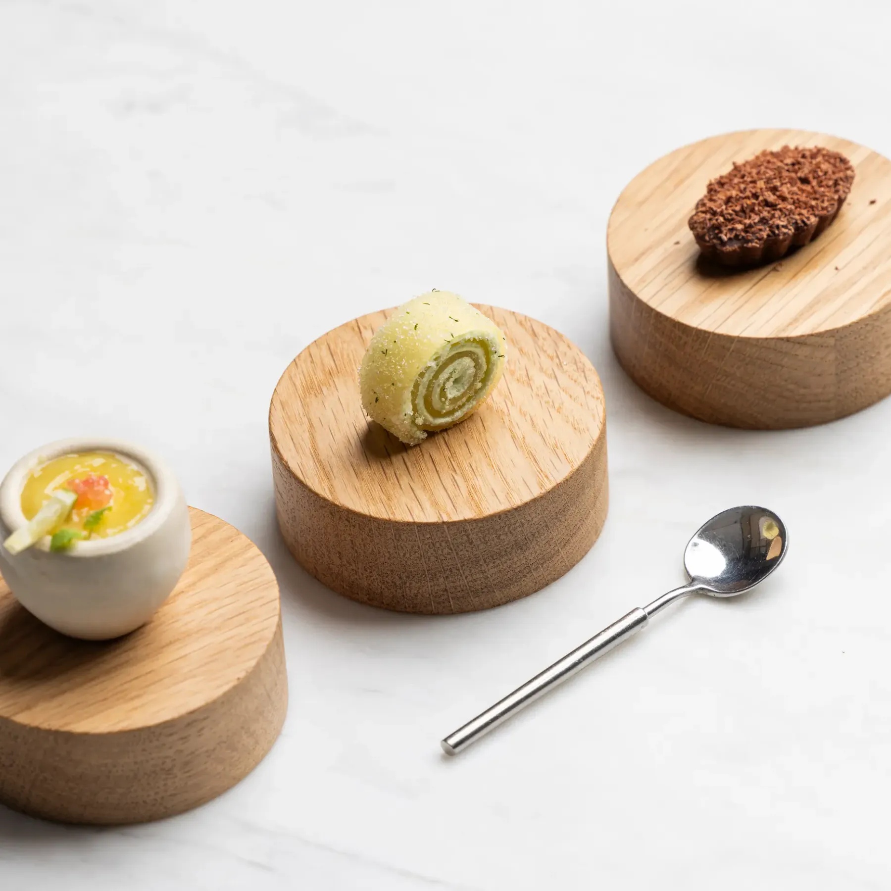 Three small portions of food on wooden stands with a spoon beside them