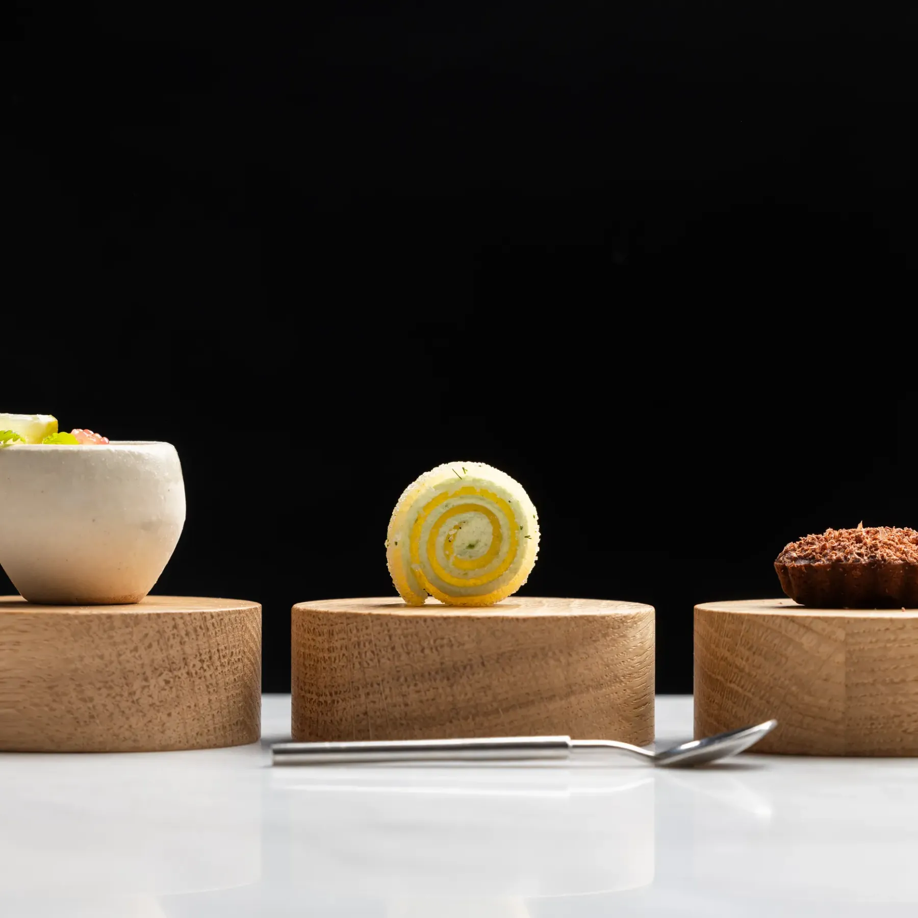 Three desserts presented on light wooden stands, including a white cup with garnish, a rolled slice, and a small cake covered in cocoa.