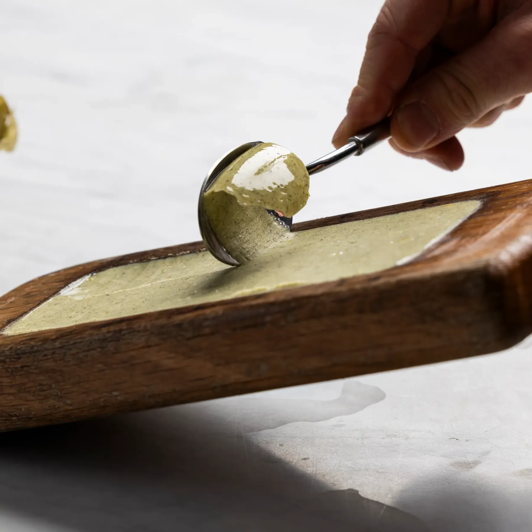 A hand spreads green dough on a wooden board using a spatula.