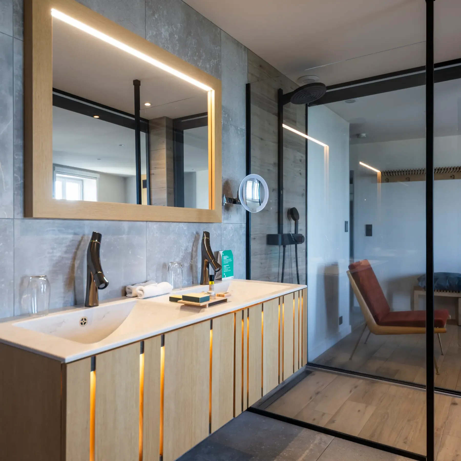 Modern bathroom with stone sink, illuminated mirror, and glass shower, overlooking an adjacent room with chair and bench.