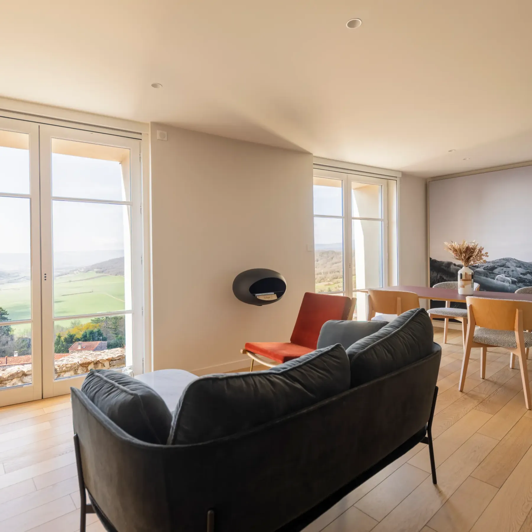 A living room with a black sofa and a wooden dining table, offering an outdoor view through large windows.