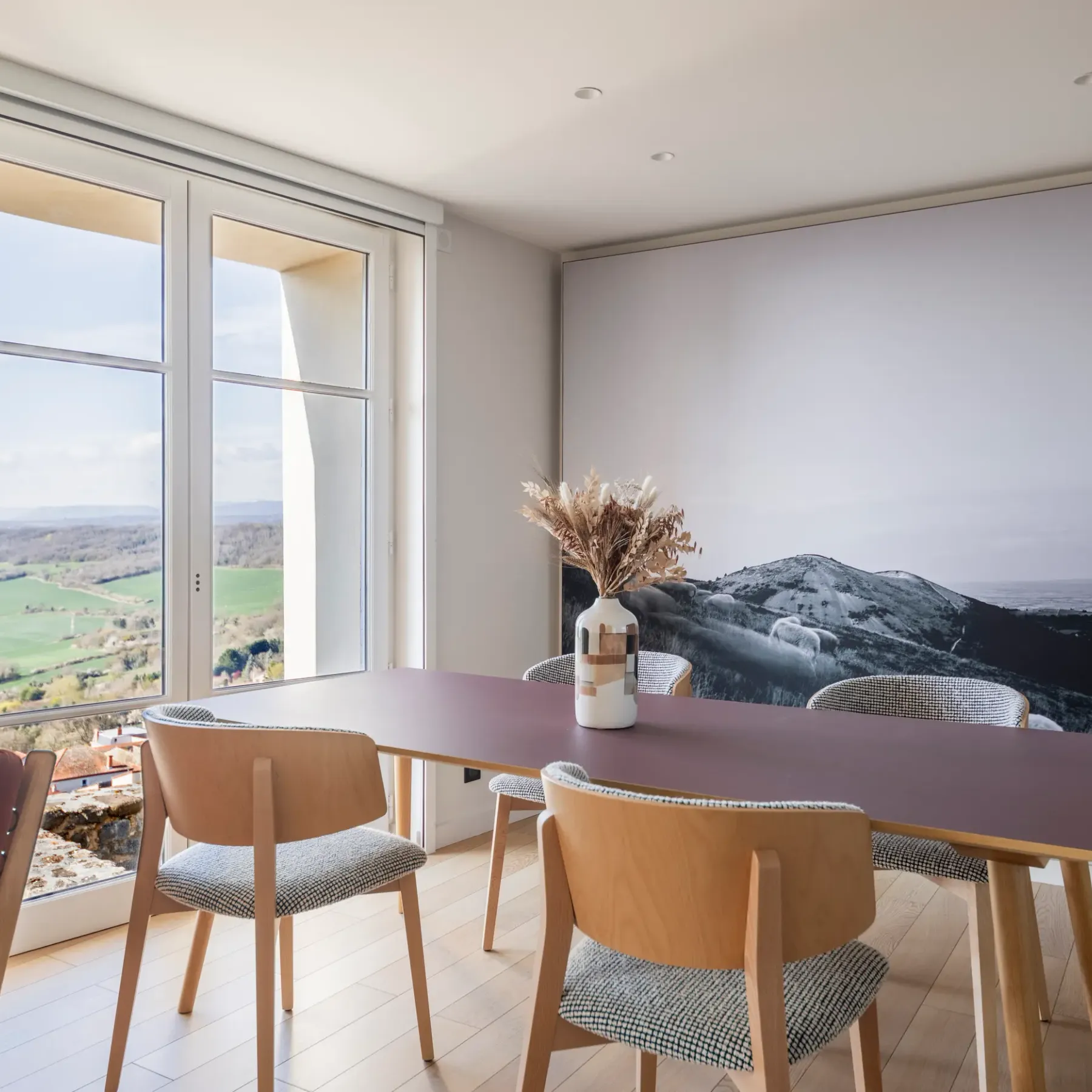 A rectangular table with chairs around it, in front of a large window offering a view of an outdoor landscape.