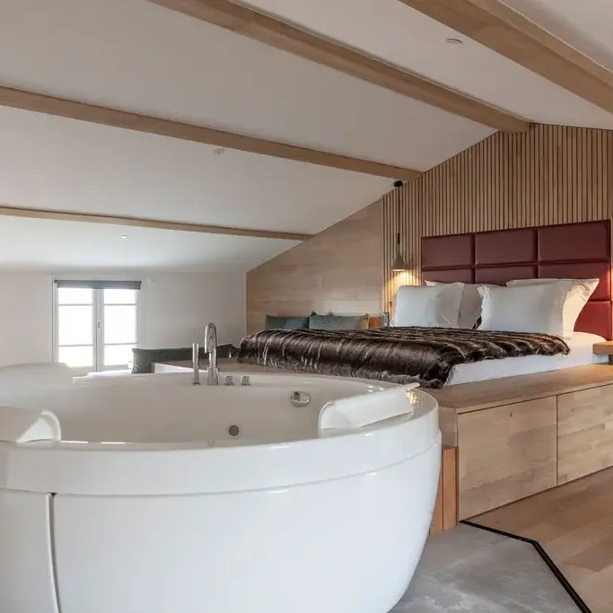 Room with a bed and a wooden headboard, a white bathtub, and windows letting in natural light.