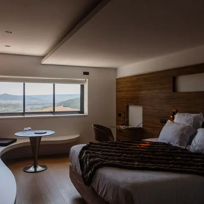 Room with bed, round table and panoramic view of mountains through large windows