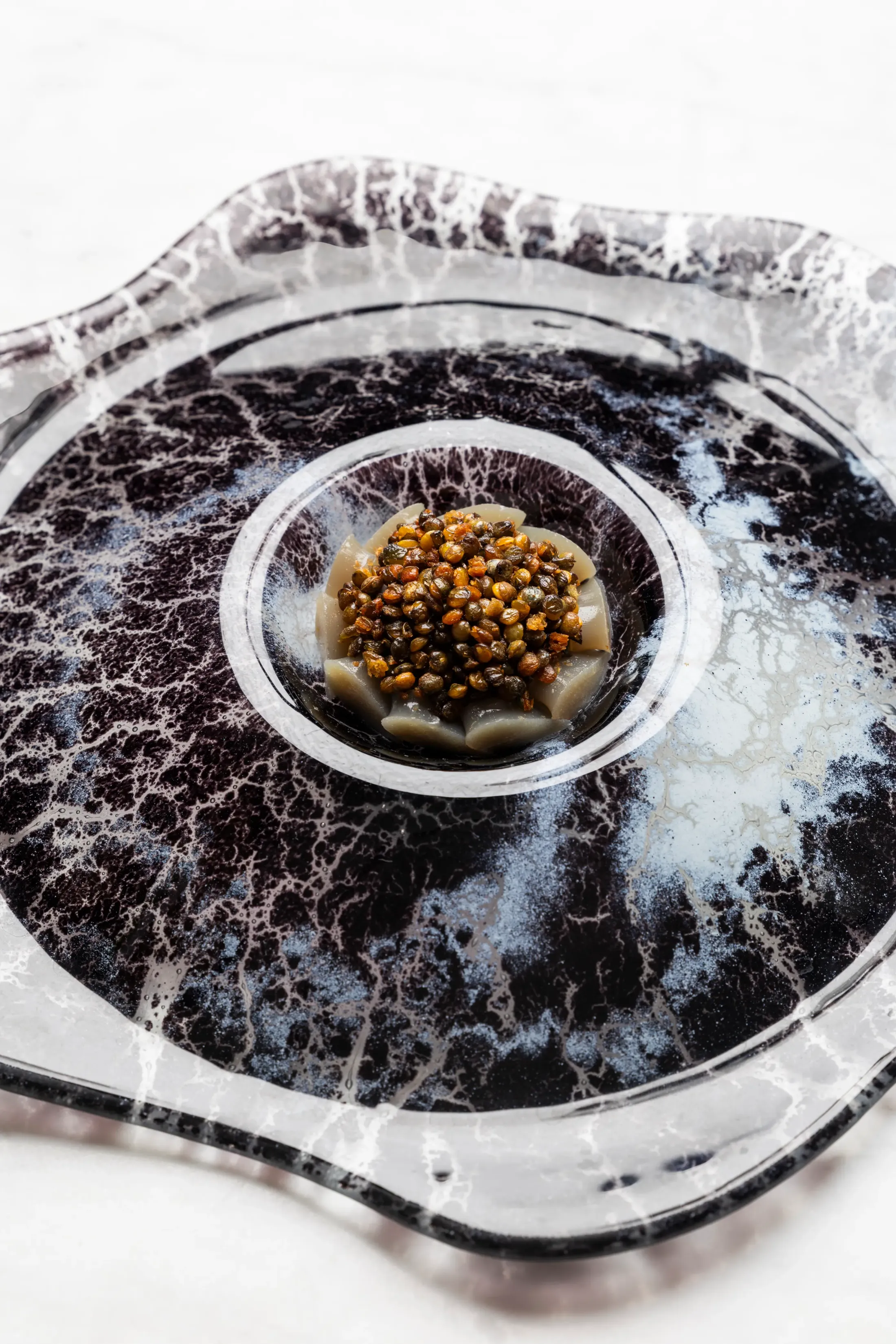 Un petit bol contenant des lentilles et des morceaux de légumes est placé au centre d'une assiette en verre marbré noir et blanc