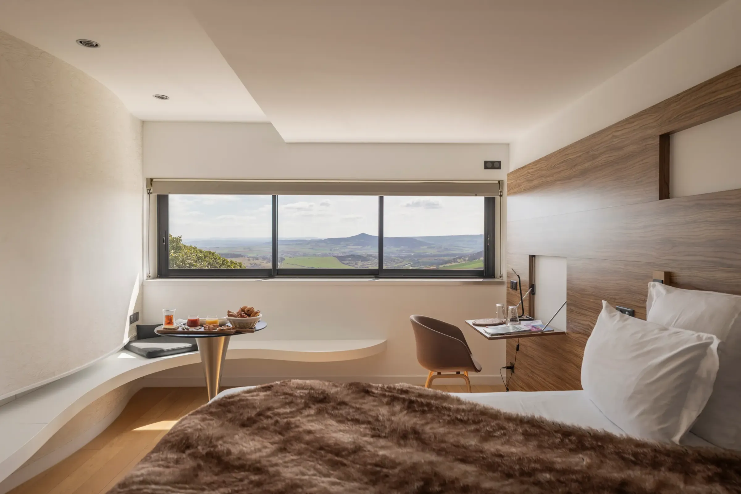Chambre avec lit, table de chevet, chaise et bureau, offrant une vue panoramique sur un paysage vallonné à travers une grande fenêtre