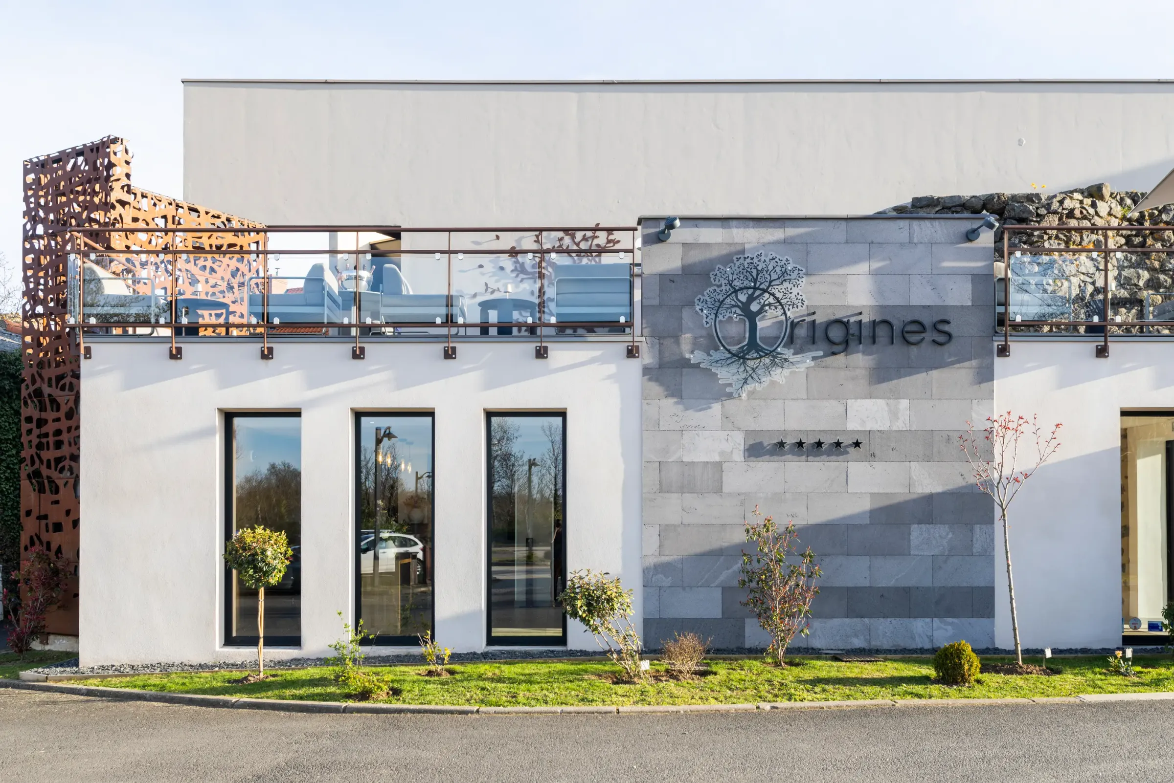 Bâtiment moderne avec façade blanche et grise, balcon à balustrade métallique et végétalisée, et enseigne "Origines" accompagnée d'un logo d'arbre