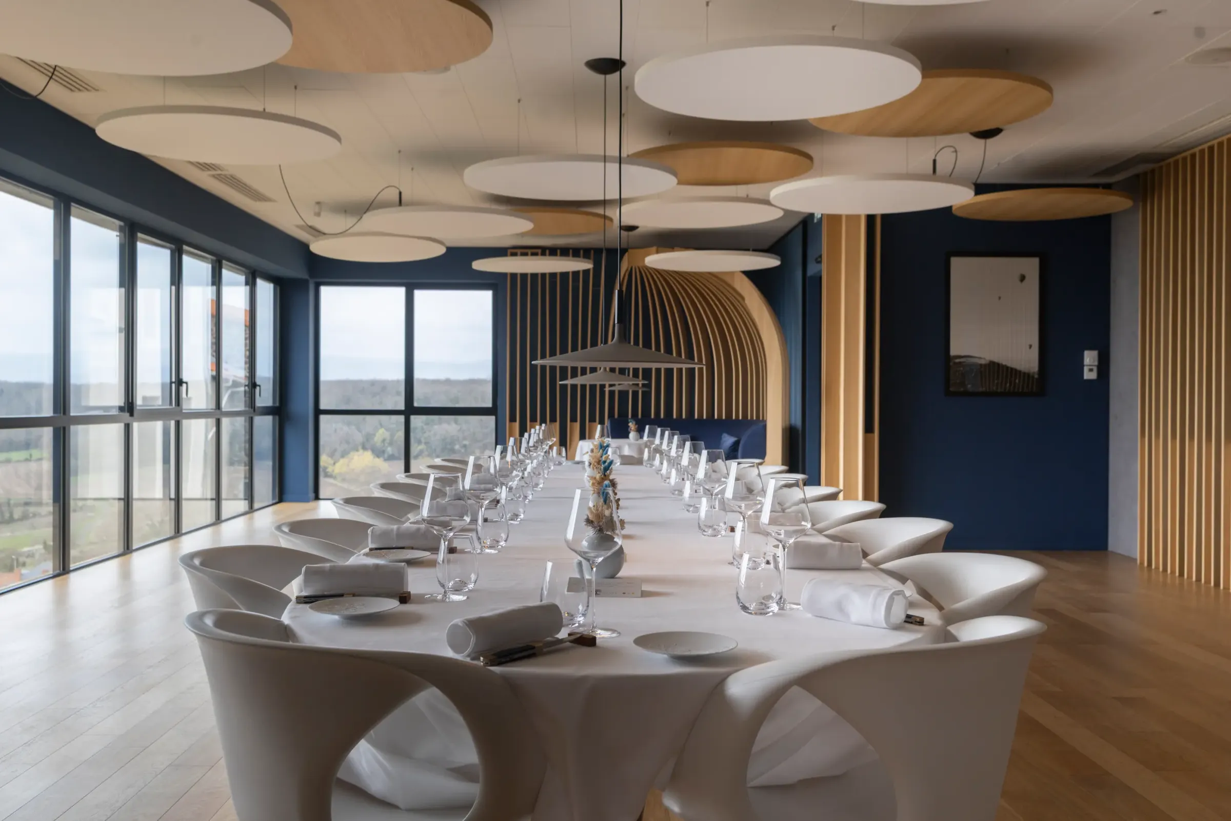 Une longue table de réunion avec des chaises modernes, des verres et des serviettes, dans une salle aux grandes fenêtres et aux luminaires circulaires suspendus