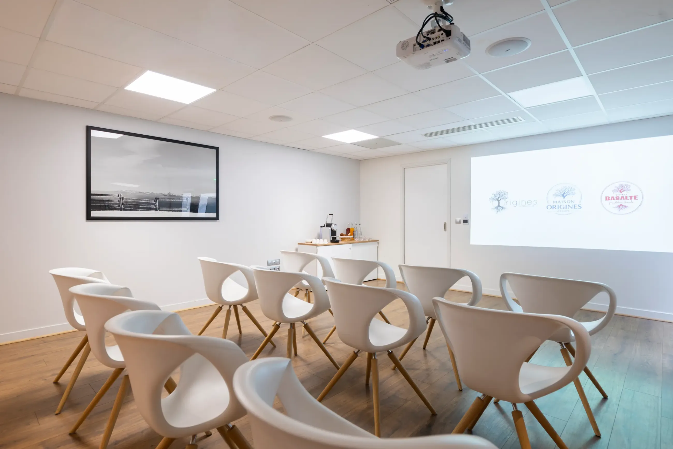 Salle de réunion avec des chaises blanches disposées en cercle, un projecteur au plafond, une photo encadrée sur le mur et des logos sur un écran