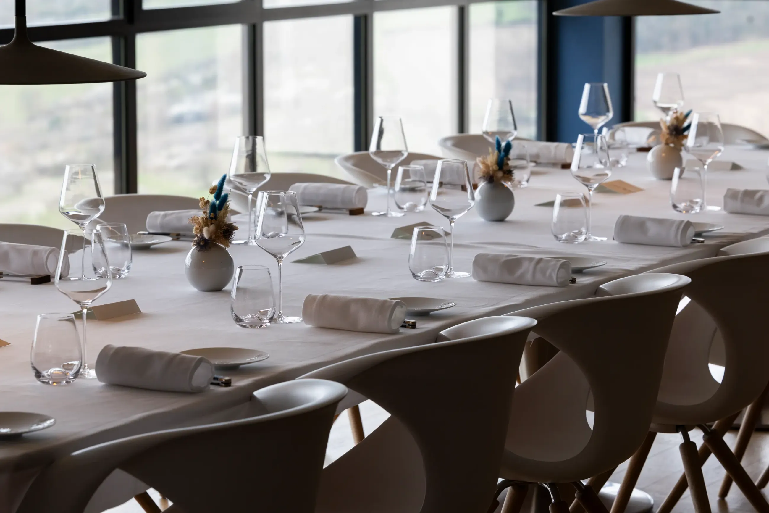 Une table de restaurant élégamment dressée avec des verres à vin, des serviettes roulées et des petits vases contenant des fleurs séchées, devant de grandes fenêtres offrant une vue extérieure