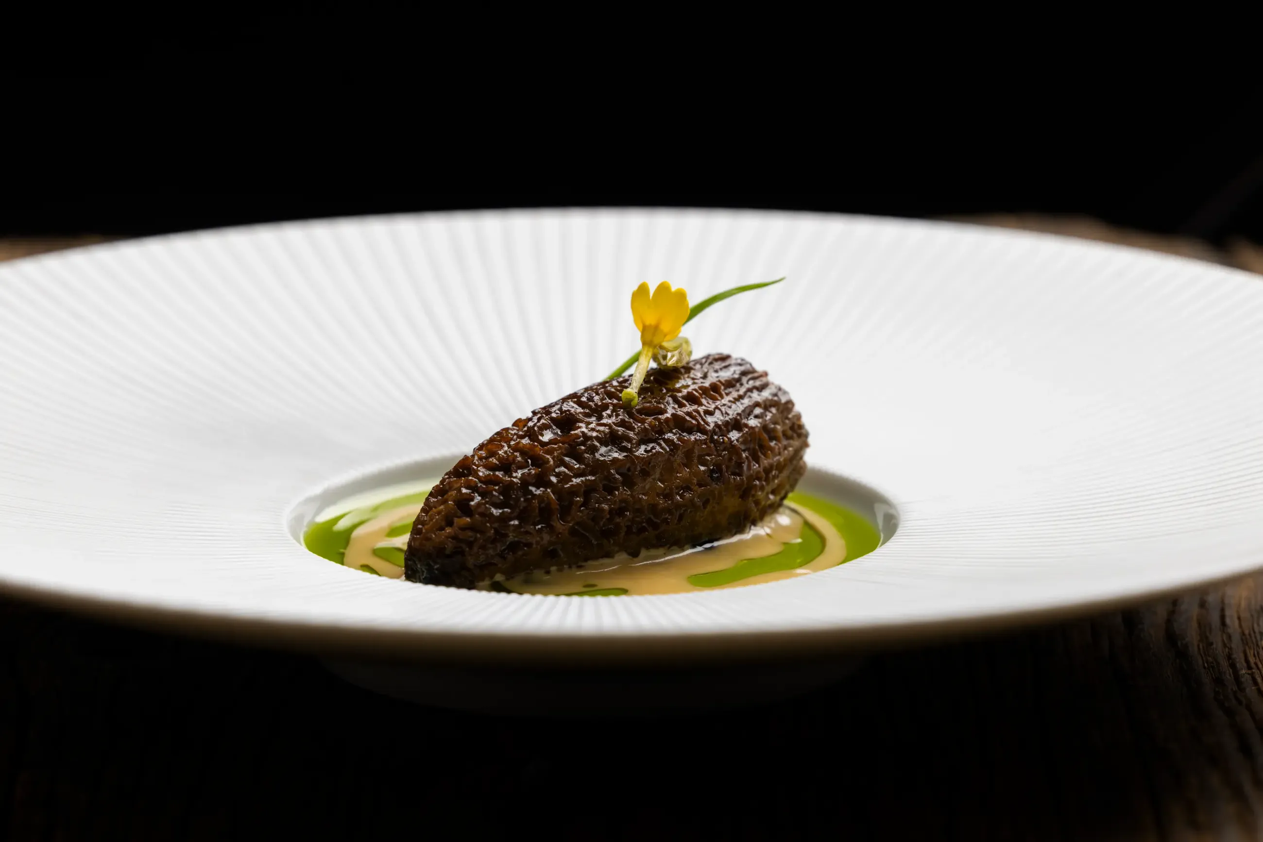 Un morceau de nourriture brun foncé est placé sur une assiette blanche avec une sauce verte autour, accompagné d'une petite fleur jaune