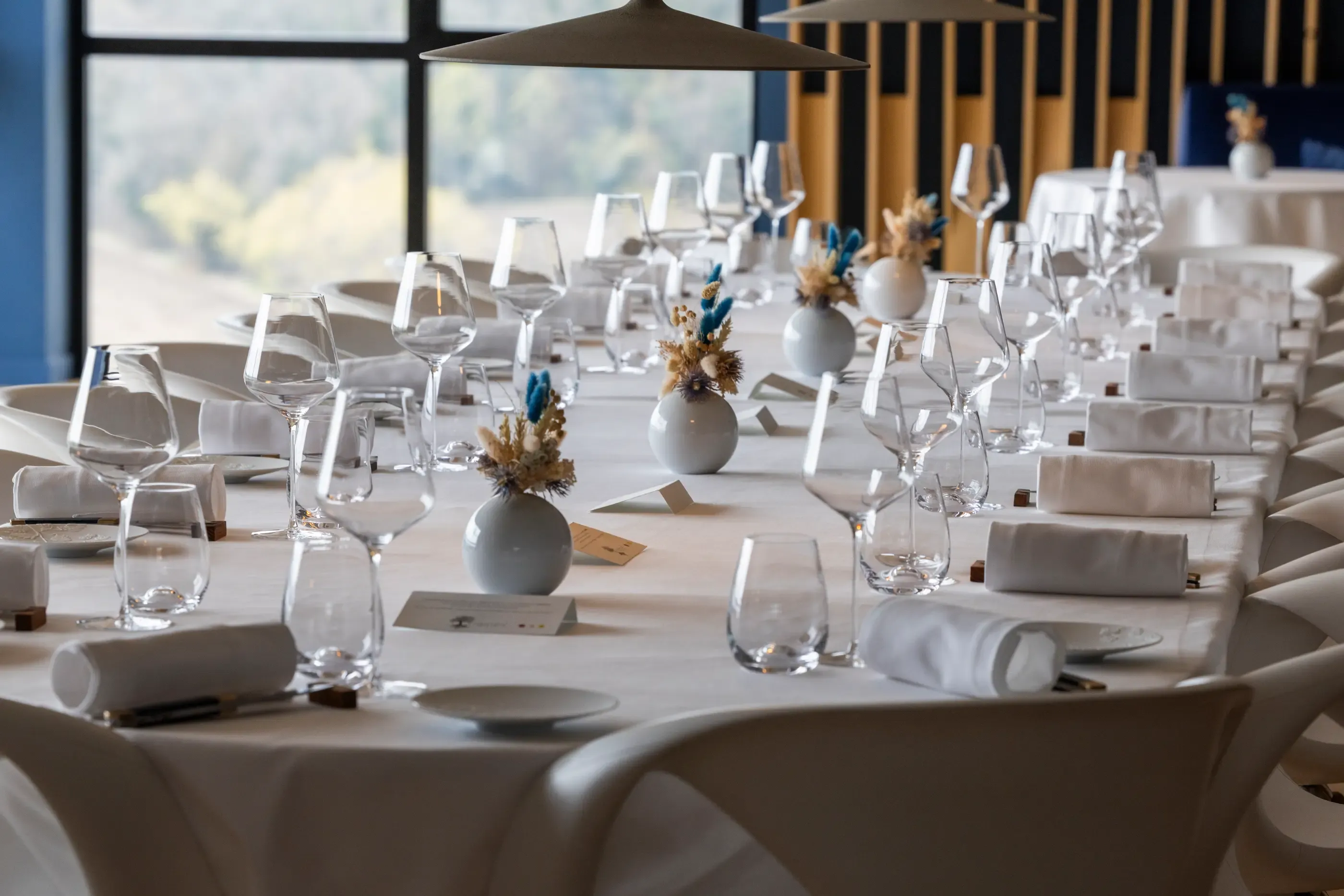 Une longue table dressée avec des verres à vin, des serviettes et des petits vases contenant des fleurs séchées