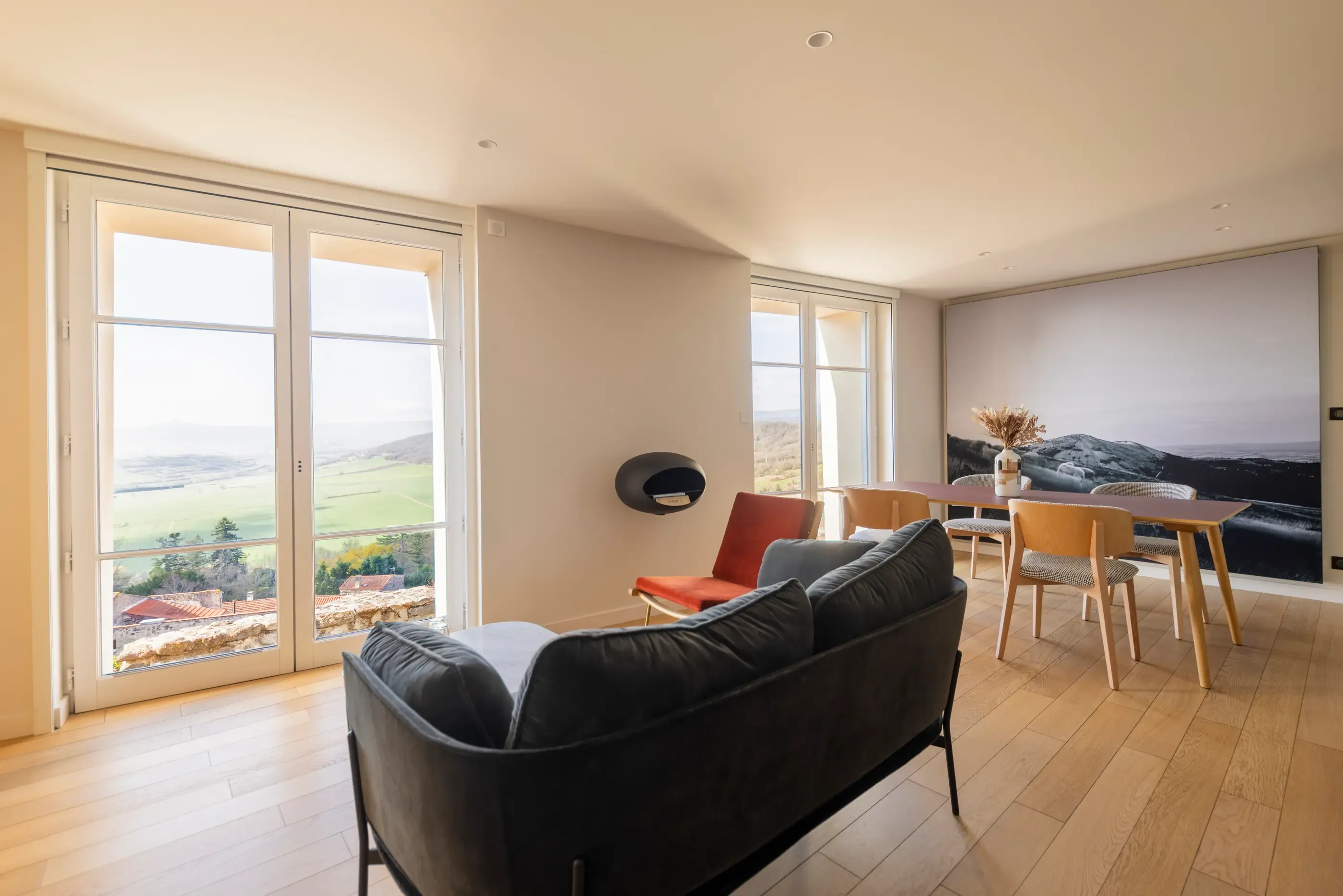 A living room with a black sofa and a wooden dining table, offering an outdoor view through large windows.