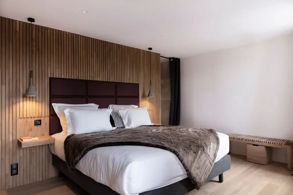Room with a double bed against a wooden wall, a gray blanket, and two lit wall lamps