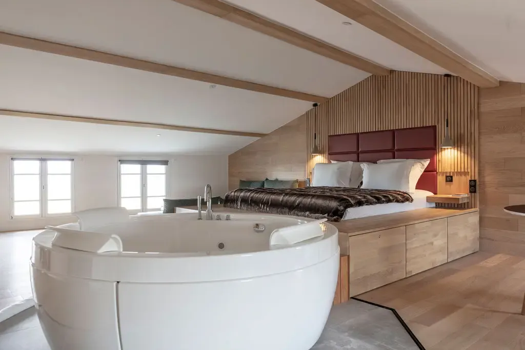 Room with a bed and a wooden headboard, a white bathtub, and windows letting in natural light.