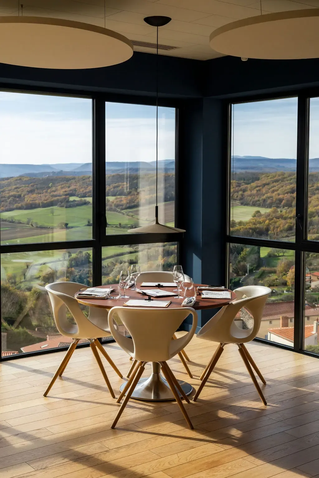 A round table with four chairs and wine glasses, located in a glass-walled room offering a panoramic view of a rolling landscape.