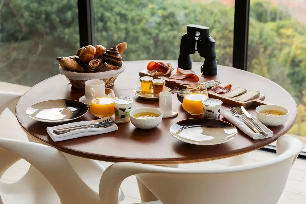 Table ronde avec un petit-déjeuner composé de pains, de charcuterie, de fromage, de boissons et d'assiettes, vue à travers une grande fenêtre donnant sur une forêt