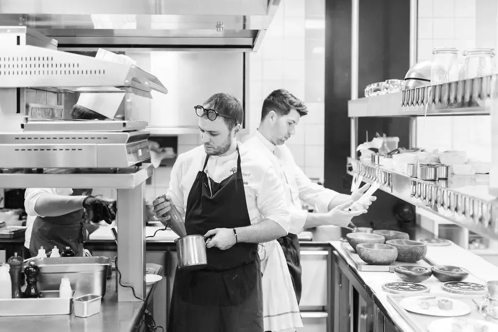 Deux cuisiniers en tablier noir préparent des plats dans une cuisine professionnelle équipée de divers ustensiles et ingrédients